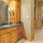 Bathroom with wood cabinets, a dark granite countertop, sink, wall-mounted light fixtures, and a large mirror. Tile floor leads to a bedroom visible through a doorway, with a bed and windows overlooking greenery.