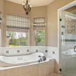 A spacious bathroom features a large white soaking tub beneath three windows with striped Roman shades, a glass-enclosed shower, beige tile flooring, and soft tan walls.