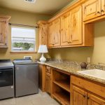 Laundry room with wood cabinets, granite countertop, sink, lamp, two front-loading machines, tile floor, and a window with a white shade. The walls are tan, and there is open shelving above the machines.