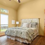 A cozy bedroom with a wooden floor, light yellow walls, a rustic wooden door, a bed with patterned bedding, a white lamp on a nightstand, and three large windows with shades letting in natural light.
