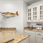 A small home bar area with cream cabinets, granite countertops, a glass-front cabinet, a mounted fish, and an antelope head sculpture on the counter. A framed photo and faucet are also visible.