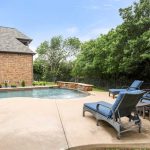 A backyard with a swimming pool, surrounded by trees and greenery. Four blue cushioned lounge chairs are arranged on the concrete pool deck, and a brick house is visible on the left. The sky is clear with a few clouds.