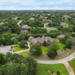Aerial view of a suburban neighborhood with large brick homes surrounded by dense green trees, curving roads, and well-kept lawns under a cloudy sky.
