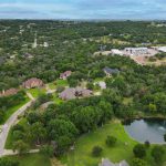 Aerial view of a suburban neighborhood with large houses surrounded by dense green trees, winding roads, a small pond, and a swimming pool, with hills and open land in the background under a cloudy sky.