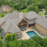 Aerial view of a large brick house with a complex roof design and a backyard pool, surrounded by green trees and lawns in a suburban neighborhood.