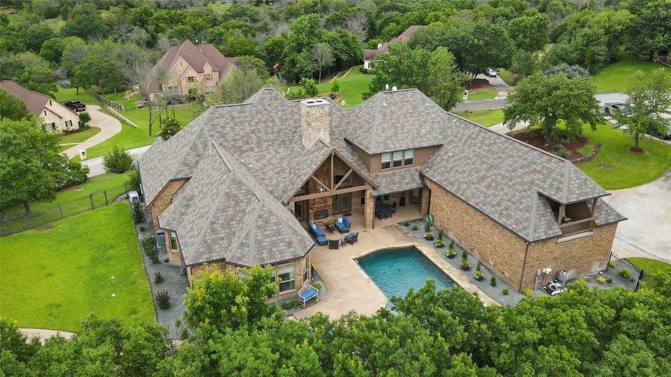 Aerial view of a large brick house with a complex roof design and a backyard pool, surrounded by green trees and lawns in a suburban neighborhood.