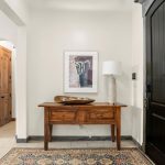 A wooden entry table with two drawers sits against a cream wall, topped with a bowl and a white lamp. A framed abstract artwork hangs above. A dark door and patterned rug are visible. An open archway leads to another room.