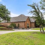 A large brick house with a steep, shingled roof sits surrounded by green grass, mature trees, and a spacious curved driveway under a partly cloudy sky.