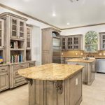 Spacious kitchen featuring wooden cabinets, glass-front upper cupboards, a central island with a marble countertop, stainless steel appliances, and beige tiled flooring. A large window above the sink lets in natural light.