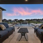 Two outdoor wicker sofas with blue cushions face each other on a covered patio, overlooking a rectangular pool and landscaped backyard at sunset with pink clouds in a blue sky.