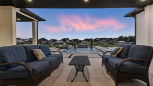 Two outdoor wicker sofas with blue cushions face each other on a covered patio, overlooking a rectangular pool and landscaped backyard at sunset with pink clouds in a blue sky.