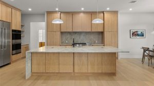 Modern kitchen with light wood cabinets, a large marble island with a built-in sink, stainless steel appliances, and two pendant lights. A dining area with a table and chairs is visible on the right.