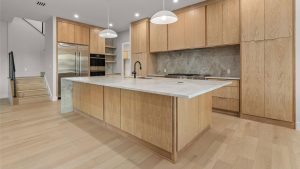 Modern kitchen with light wood cabinets, a large center island with a marble countertop, built-in stainless steel appliances, pendant lights, and light wood flooring. Stairs are visible on the left side.