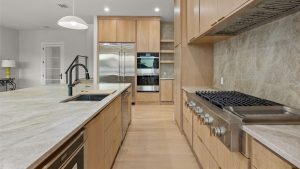 Modern kitchen with light wood cabinets, stainless steel appliances including a double oven and refrigerator, marble countertops, a built-in gas stove, and a large island with a sink and black faucet.