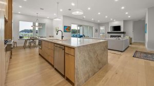 Modern open-concept kitchen and living room with light wood floors, large marble island, pendant lights, neutral-colored furniture, wall-mounted TV, and large windows letting in natural light.