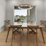 A modern dining room with a wooden table and four chairs, a round chandelier overhead, framed artwork on the walls, and a large window showing a lakeside view with houses outside.