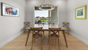A modern dining room with a wooden table and four chairs, a round chandelier overhead, framed artwork on the walls, and a large window showing a lakeside view with houses outside.