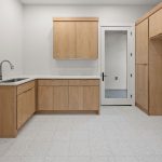 A minimalist kitchen with light wood cabinets, white countertops, a stainless steel sink, and a white tile floor. A glass door leads to another room, and the walls are painted light gray.