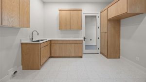 A minimalist kitchen with light wood cabinets, white countertops, a stainless steel sink, and a white tile floor. A glass door leads to another room, and the walls are painted light gray.