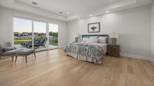 A spacious bedroom with light wood floors, a large bed with patterned bedding, two nightstands with lamps, a lounge chair, and sliding glass doors opening to a patio with lounge chairs and a grassy yard.