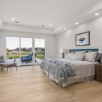 A spacious, modern bedroom with light wood floors, a large bed with patterned bedding, two wooden nightstands, and a gray chair by the glass doors that open to a patio with a view of a backyard and neighboring houses.