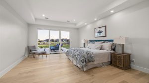 A spacious, modern bedroom with light wood floors, a large bed with patterned bedding, two wooden nightstands, and a gray chair by the glass doors that open to a patio with a view of a backyard and neighboring houses.