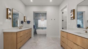 Modern bathroom with double vanities featuring wood cabinets and large mirrors, white countertops, gold fixtures, and a central freestanding tub set against blue marble walls, with sconces and recessed lighting.