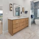 Modern bathroom with a wooden vanity, double sinks, large mirror, and gold sconces; a bathtub sits in a tiled alcove with a chandelier, and the room connects to a bedroom with large windows overlooking a backyard.