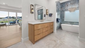 Modern bathroom with a wooden vanity, double sinks, large mirror, and gold sconces; a bathtub sits in a tiled alcove with a chandelier, and the room connects to a bedroom with large windows overlooking a backyard.