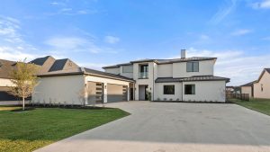 Modern two-story house with light-colored exterior, large windows, and a three-car garage. The house has a wide driveway, manicured lawn, and is set against a blue sky with wispy clouds.