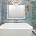 Luxurious bathroom with a white freestanding tub, blue-green marble walls, a frosted window, three blue towels, a gold faucet, glass shower enclosure, and a modern chandelier overhead.