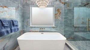 Luxurious bathroom with a white freestanding tub, blue-green marble walls, a frosted window, three blue towels, a gold faucet, glass shower enclosure, and a modern chandelier overhead.