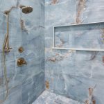 Modern shower with blue and white marble-patterned tiles, brass shower fixtures, a rainfall showerhead, a handheld sprayer, and a built-in niche shelf inset into the wall. The floor features small hexagonal tiles.
