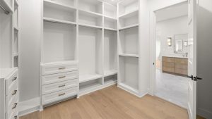 Spacious walk-in closet with white built-in shelves, drawers, and hanging rods, featuring light wood flooring. An open doorway reveals a modern bathroom with a double vanity and mirrors.