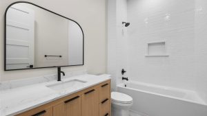 Modern bathroom with a wooden vanity, white countertop, black fixtures, large mirror, white toilet, and a white tiled bathtub-shower combo with built-in shelf.