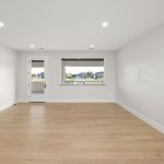 A spacious, empty room with light wood flooring, white walls, recessed ceiling lights, a large window with an outdoor view, and a door leading to a balcony or patio.