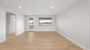 A spacious, empty room with light wood flooring, white walls, recessed ceiling lights, a large window with an outdoor view, and a door leading to a balcony or patio.