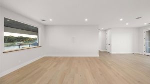 A bright, empty room with light wood floors, white walls, recessed ceiling lights, a large window overlooking a river, and French doors leading to another area.