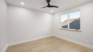 A bright, empty room with light wood floors, white walls, a modern black ceiling fan, recessed lighting, and a window showing rooftops outside.