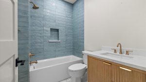 Modern bathroom with a bathtub and shower, blue tiled wall, built-in shelf, white toilet, white countertop, wood vanity, gold fixtures, and white walls.
