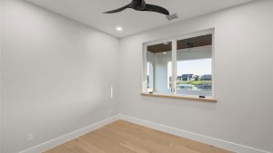 A small, empty room with light wood flooring, white walls, a black ceiling fan, recessed ceiling lights, and a large window overlooking houses and a body of water.
