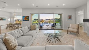 A modern living room with a light gray sofa, glass coffee table, and rug, opening onto a patio with a pool and outdoor dining area. Large sliding glass doors provide a view of houses and greenery outside.