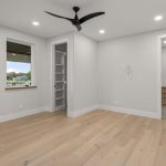 A modern, empty bedroom with light wood flooring, white walls, a ceiling fan, a small window, open closet shelves, and a view into a bathroom with light wood cabinetry. Recessed lights illuminate the space.