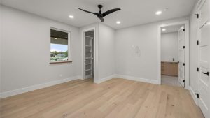 A modern, empty bedroom with light wood flooring, white walls, a ceiling fan, a small window, open closet shelves, and a view into a bathroom with light wood cabinetry. Recessed lights illuminate the space.