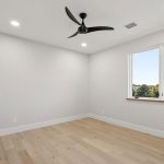 Minimalist, unfurnished bedroom with light wood flooring, white walls, a black ceiling fan, recessed lighting, a large window with a view of trees, and a white door.