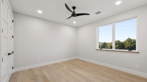 Minimalist, unfurnished bedroom with light wood flooring, white walls, a black ceiling fan, recessed lighting, a large window with a view of trees, and a white door.