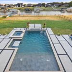 A modern backyard features a rectangular pool with an attached hot tub, surrounded by geometric concrete patio, blue lounge chairs, manicured lawn, and a view of houses and docks by a lakeside.