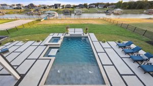 A modern backyard features a rectangular pool with an attached hot tub, surrounded by geometric concrete patio, blue lounge chairs, manicured lawn, and a view of houses and docks by a lakeside.