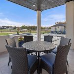 A covered patio with a round table and six wicker chairs overlooks a backyard with a swimming pool, lounge chairs, green lawn, and neighboring houses under a blue sky.