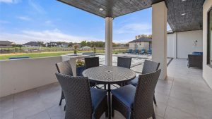 A covered patio with a round table and six wicker chairs overlooks a backyard with a swimming pool, lounge chairs, green lawn, and neighboring houses under a blue sky.